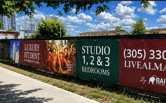 Custom printed construction fence banner installed on a chain-link fence in South Florida featuring bold black and yellow branding for DNA Marketing & Printing. The outdoor banner advertises custom construction banners available in 3–5 business days and highlights materials including 13oz vinyl banners, mesh banners, and aluminum face banners. Large-format graphics displayed at a construction site in Hallandale Beach with palm trees and blue sky in the background.